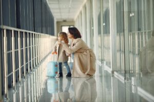 Mother crouching beside her young daughter in an airport corridor, both looking out the window with a child's suitcase nearby