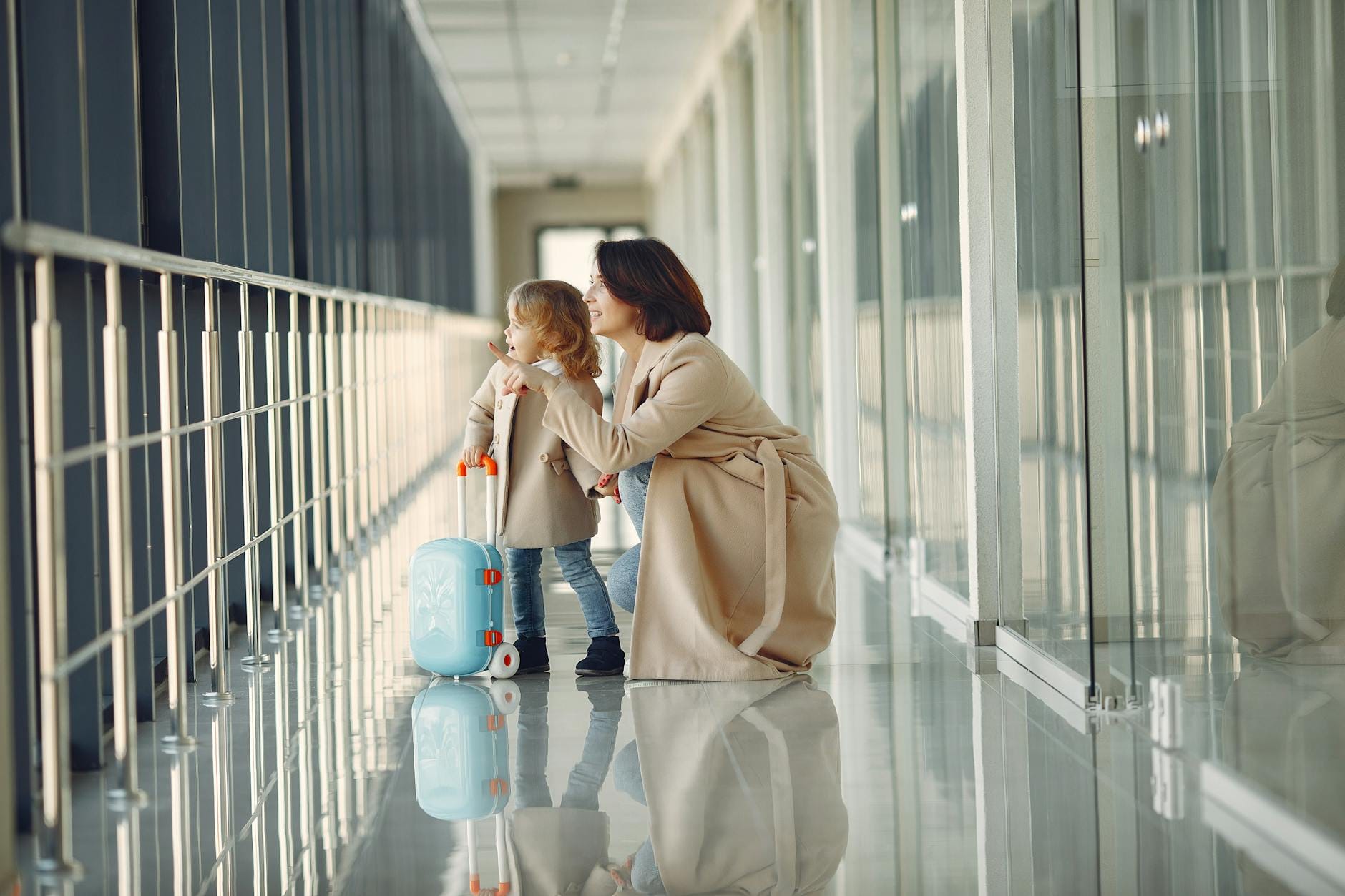 Mother crouching beside her young daughter in an airport corridor, both looking out the window with a child's suitcase nearby