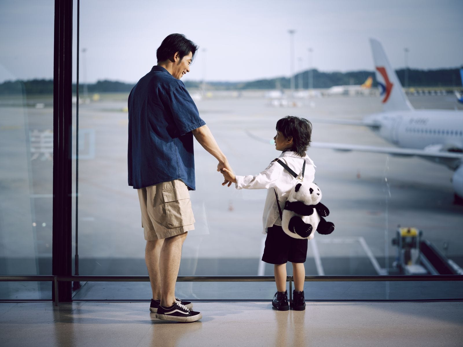 A father and child hold hands at an airport window watching planes