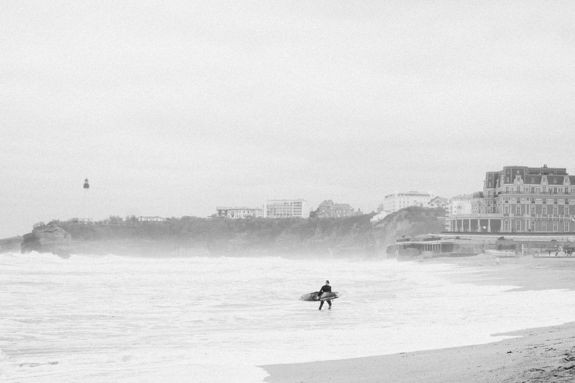 A surfer walking along Biarritz beach with the lighthouse in the background