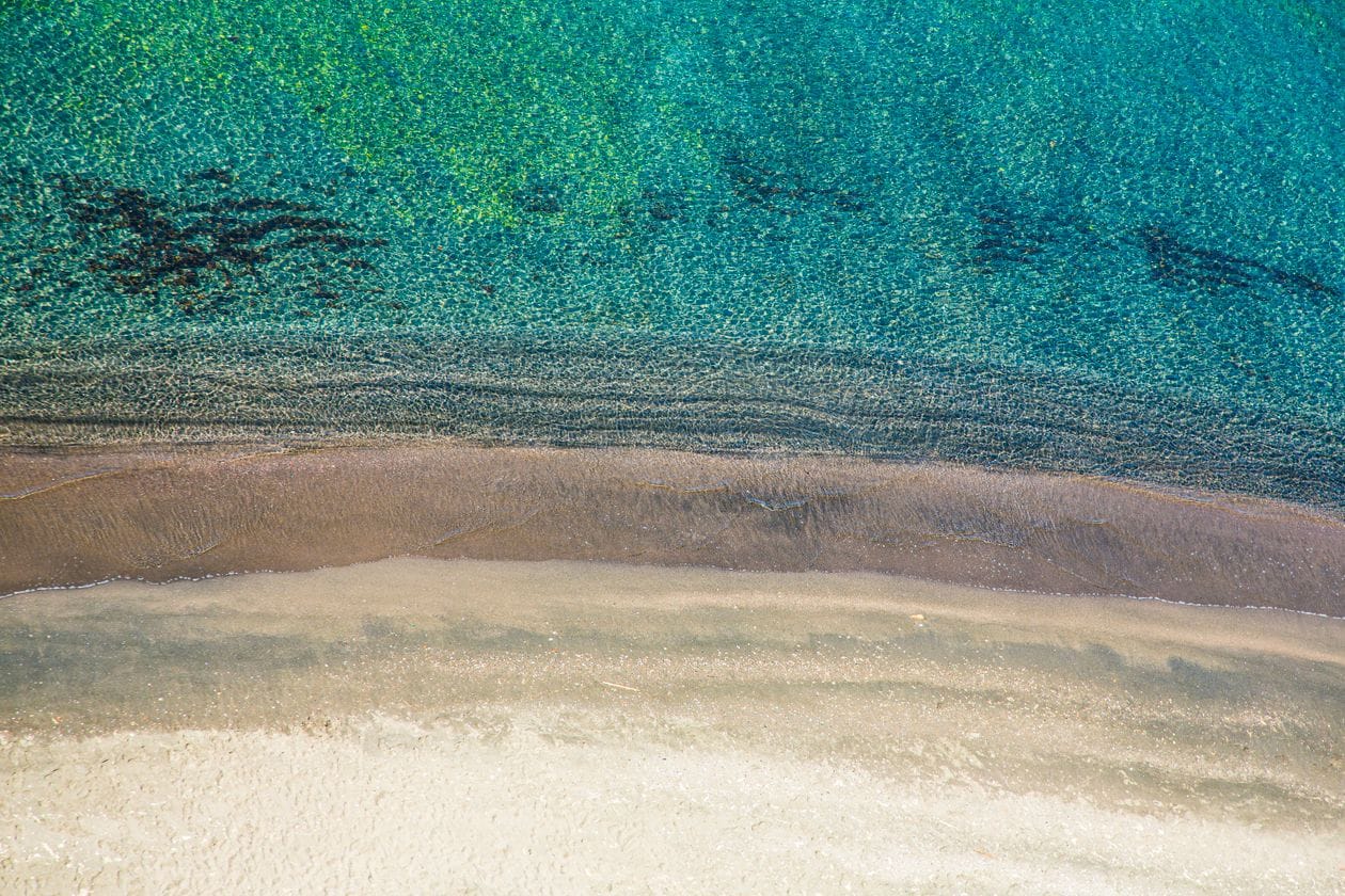 An empty stretch of white sand meeting shallow turquoise water, seen from above