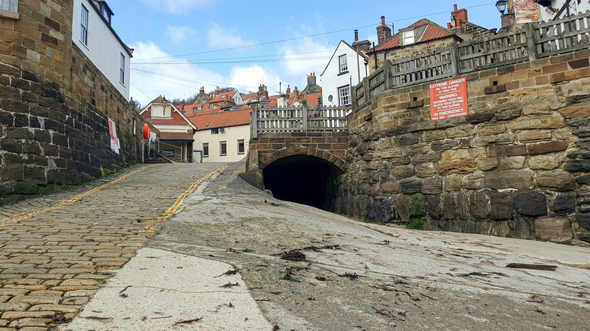 Cobbled slipway in Robin Hood's Bay leading down to the tidal sea wall, with an incoming-tide warning sign