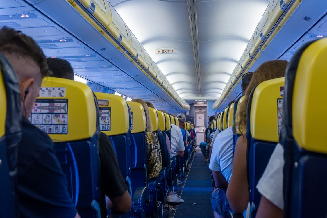 Passengers seated inside a Ryanair aircraft cabin with yellow seatbacks and an exit row sign visible