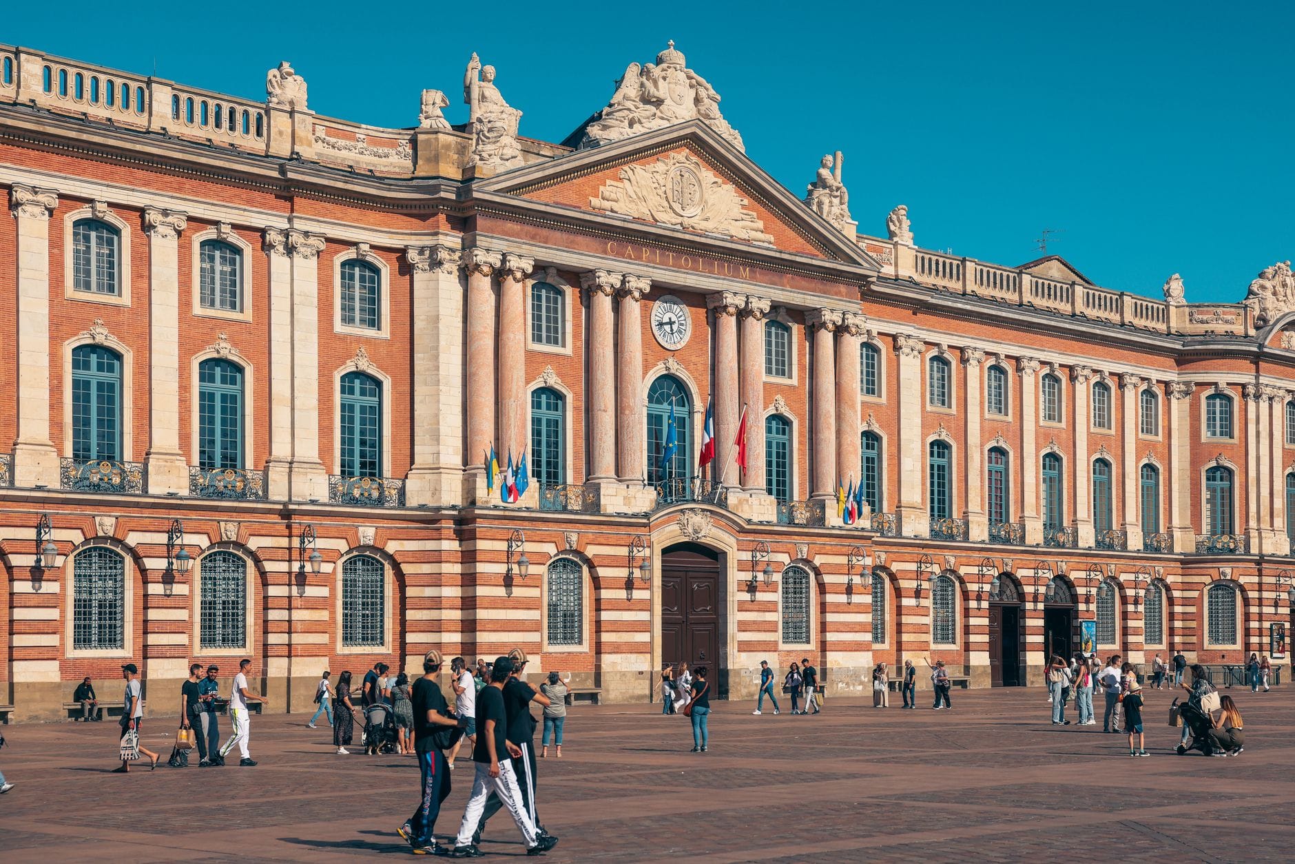 The pink-brick facade of the Capitole de Toulouse on a sunny afternoon, with visitors crossing the square