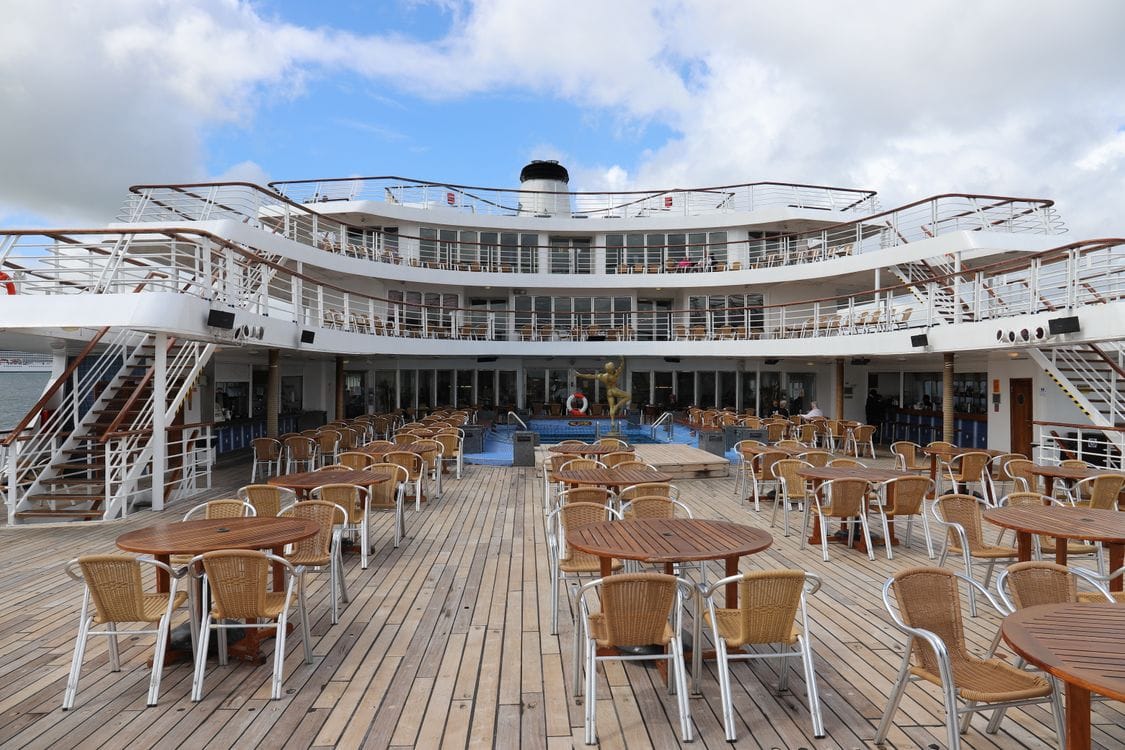 An empty cruise ship deck with tables, chairs and a pool, overlooking the sea