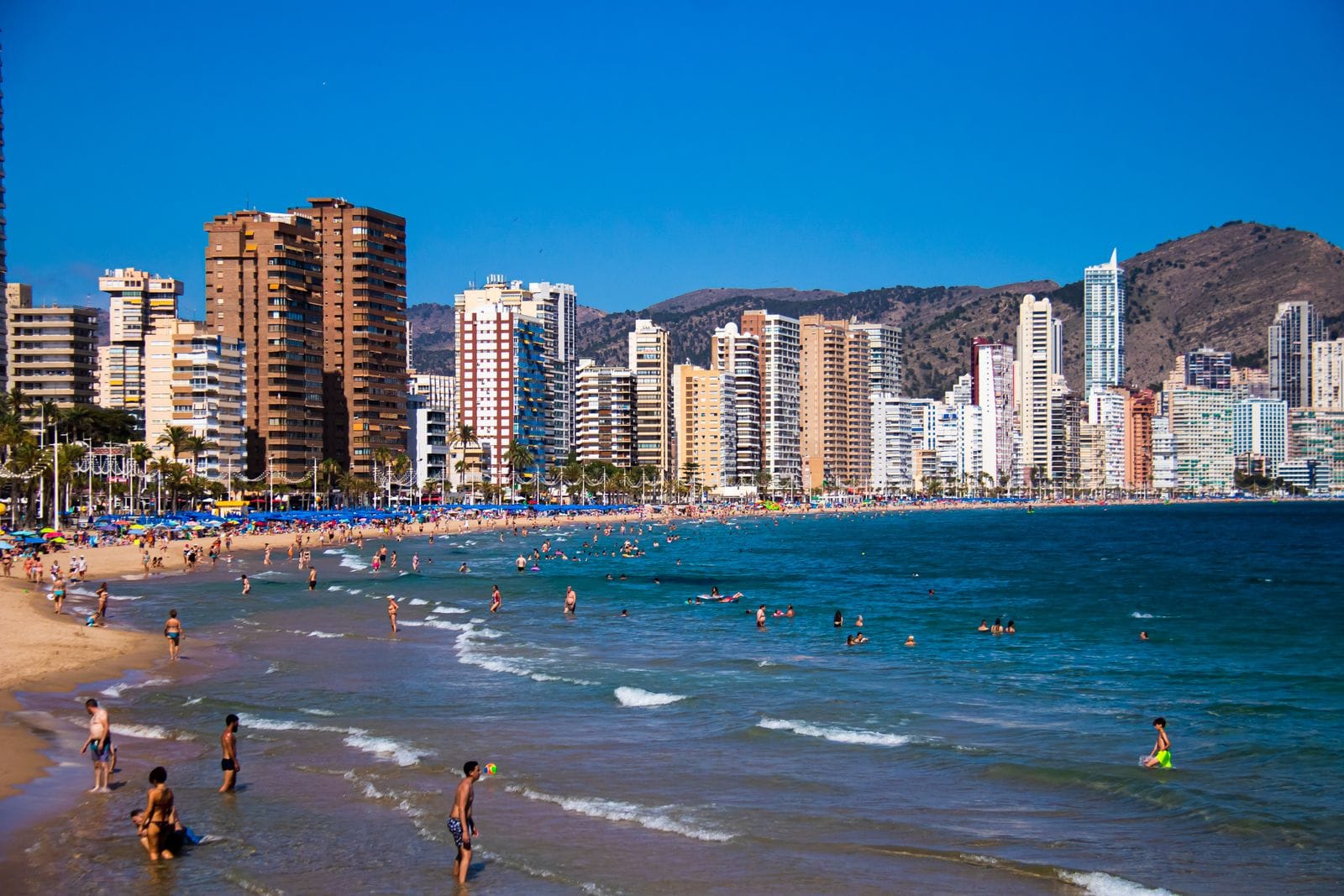 Bathers in the shallows at Benidorm's Levante beach on a busy summer day, with apartment towers rising behind the sand