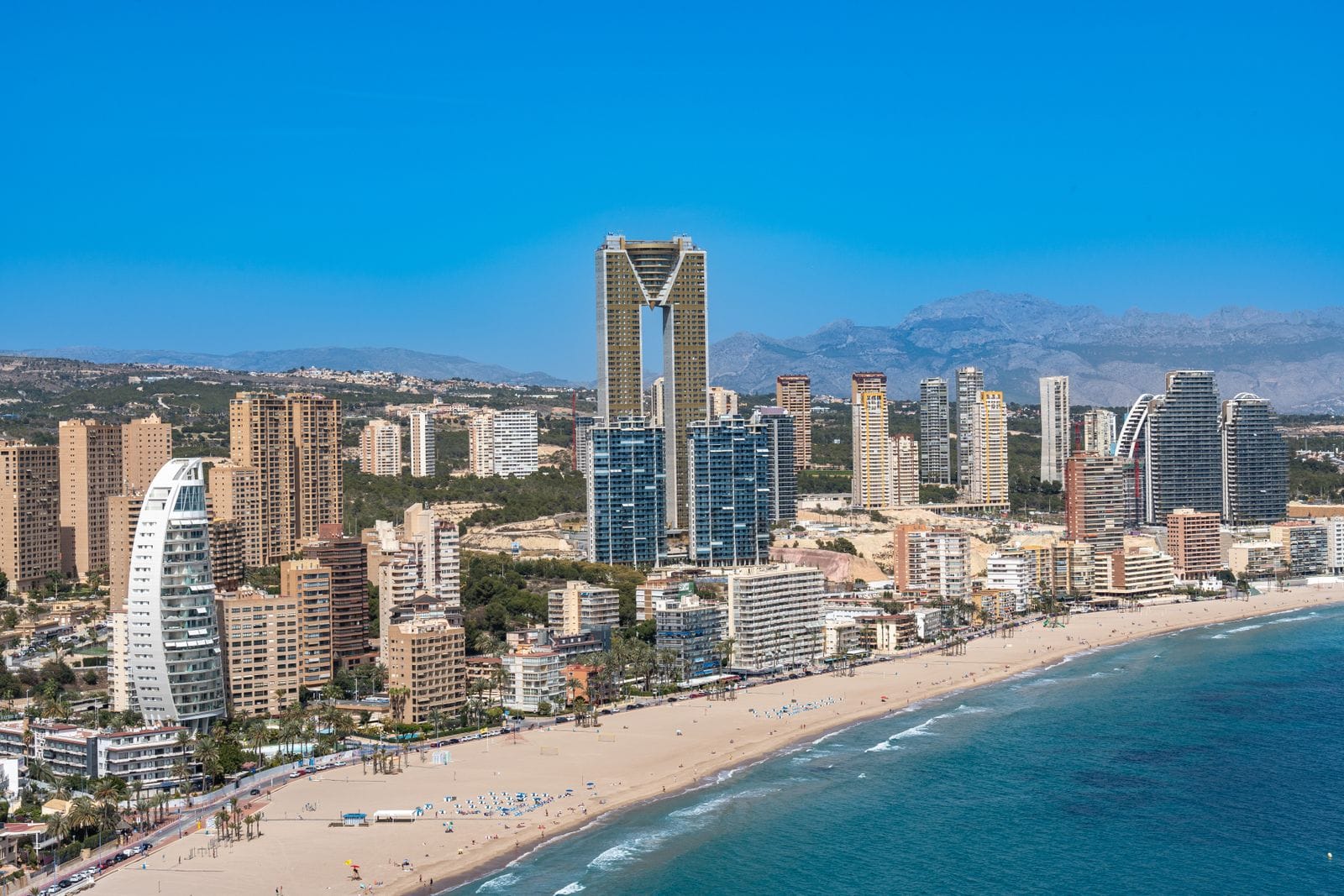Is Benidorm expensive: an aerial view of Benidorm's beachfront, with the curve of the sand and the city's cluster of high-rise towers behind it
