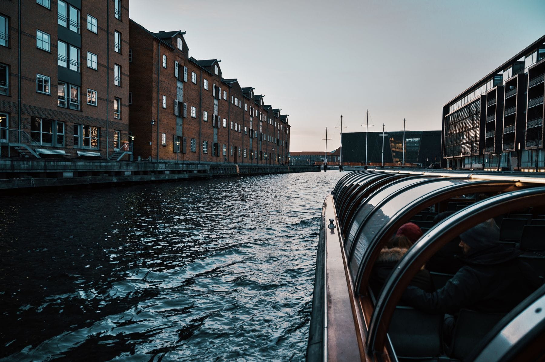 A harbour boat travelling past warehouse buildings on a Copenhagen canal, with passengers seated inside