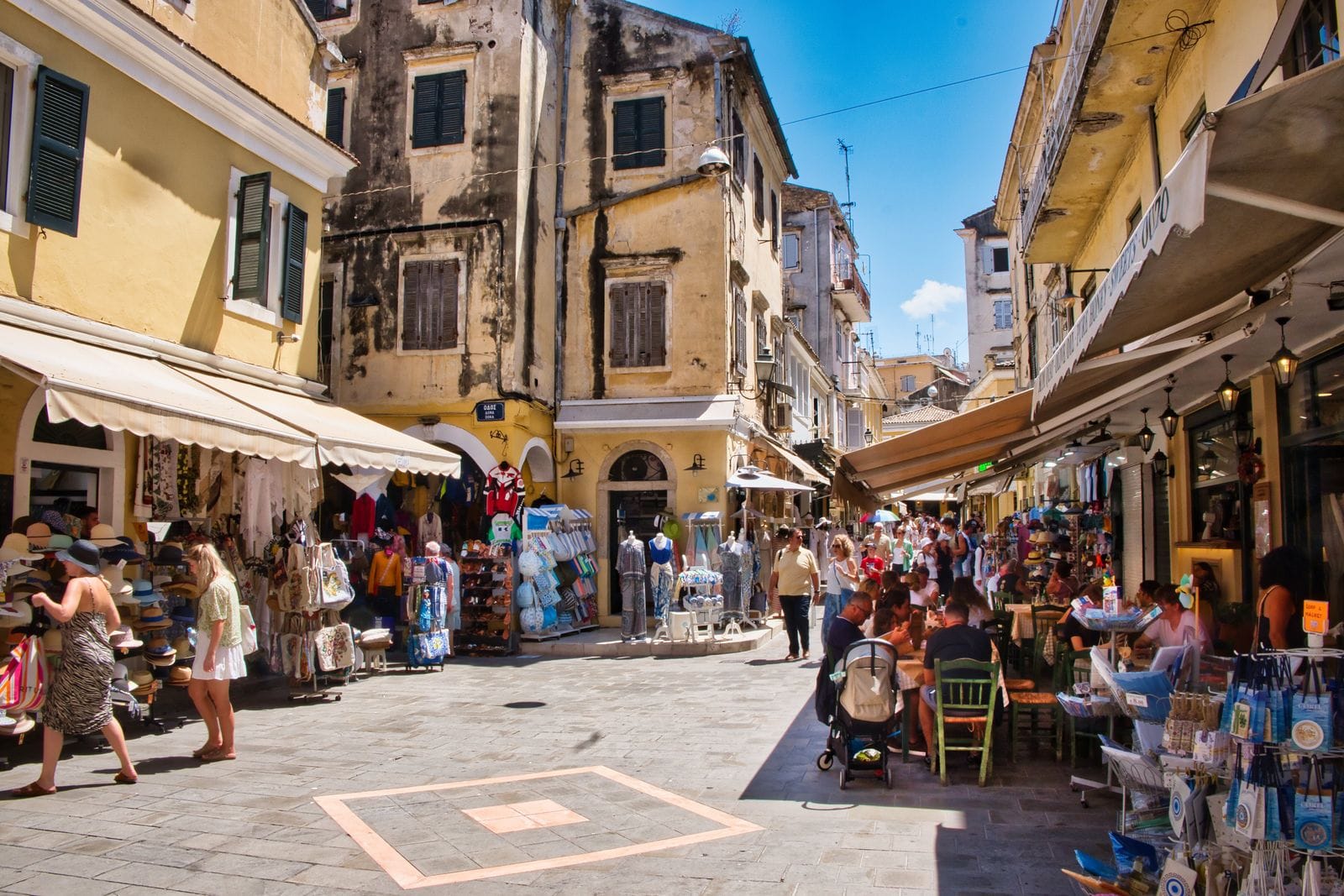 A narrow Corfu Town lane lined with awnings, souvenir stalls and cafe tables full of people