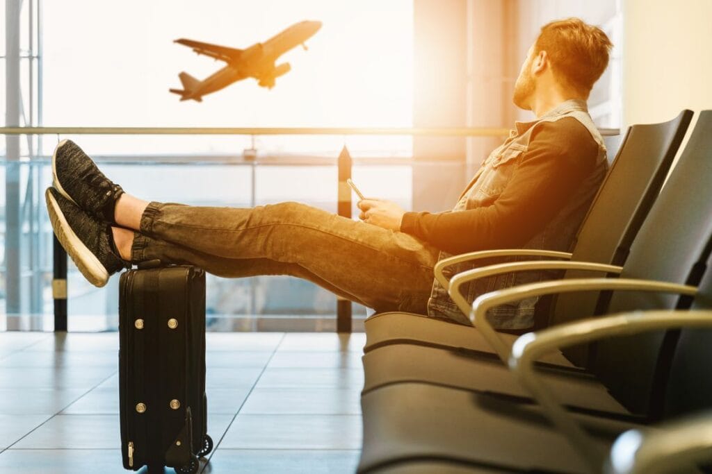 EU hand luggage rules 2027: a traveller waiting at the gate with feet resting on a rolling cabin case as a plane takes off outside