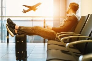 EU hand luggage rules 2027: a traveller waiting at the gate with feet resting on a rolling cabin case as a plane takes off outside