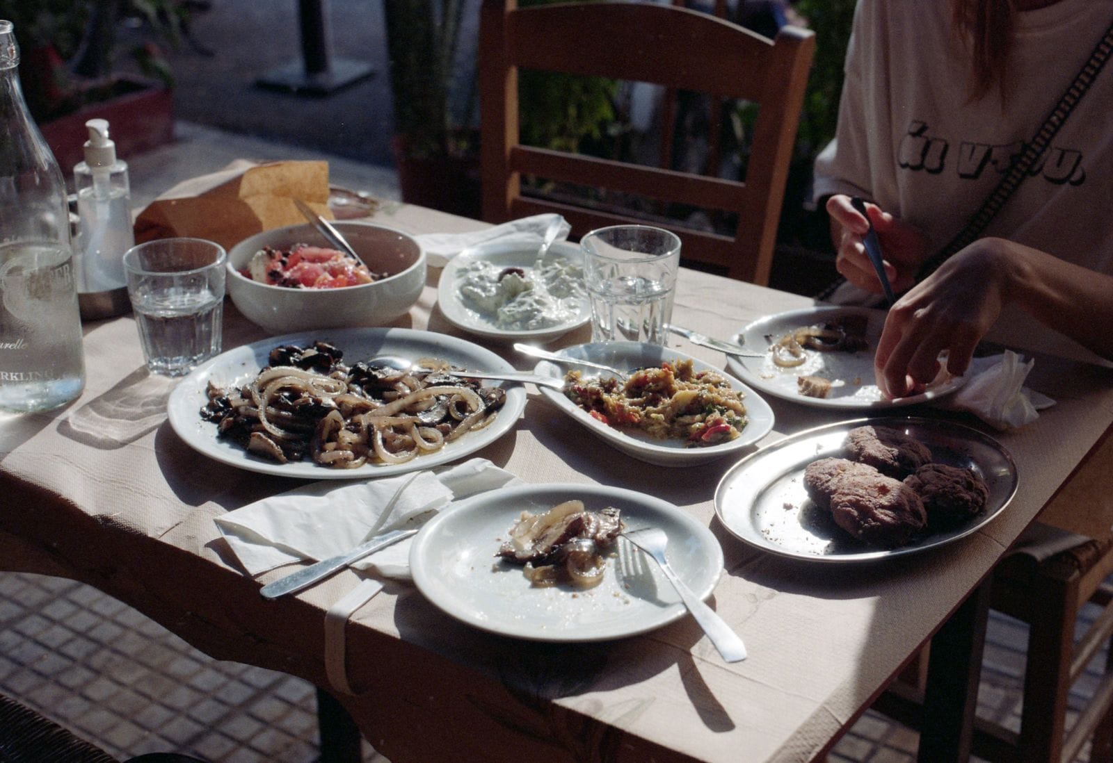 A taverna table part way through a meal, with shared plates, bread, water and wine glasses