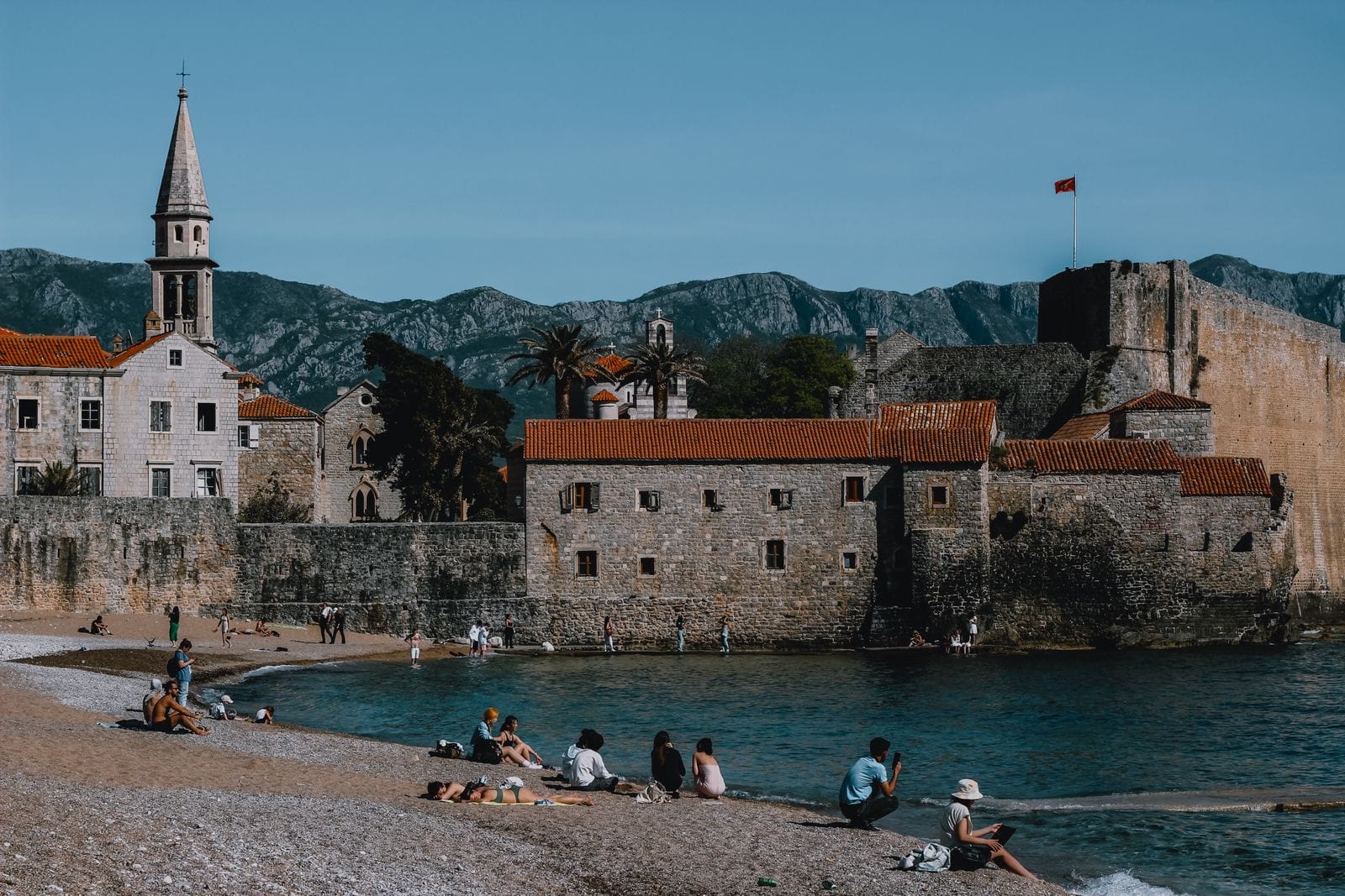 Sunbathers on the pebble beach beside the medieval stone walls of Budva old town, with mountains behind