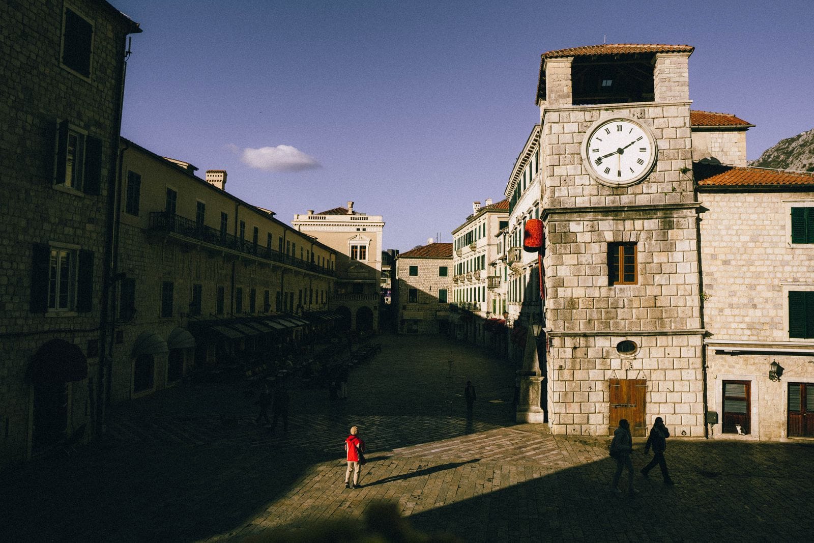 The clock tower and stone buildings of Kotor old town square at dusk, with people crossing the square