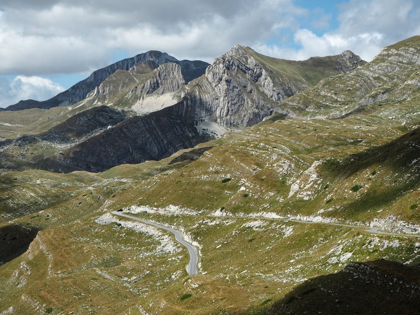 A narrow road winding through steep green mountain pasture in northern Montenegro under a cloudy sky