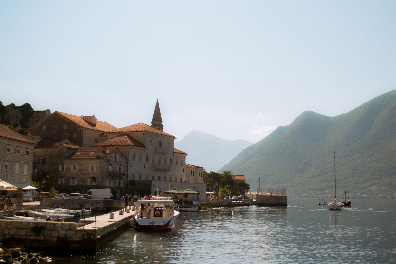 The stone waterfront of Perast on the Bay of Kotor with small boats moored, a bell tower behind and mountains across the water