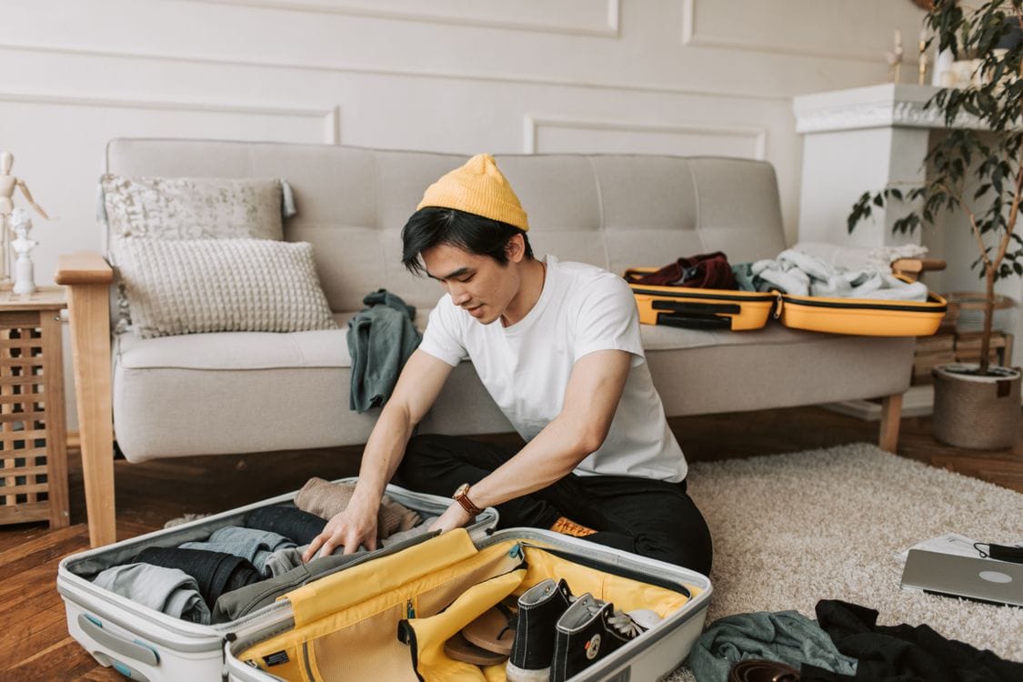 A man kneeling on a living room floor packing a hard-shell suitcase before a trip