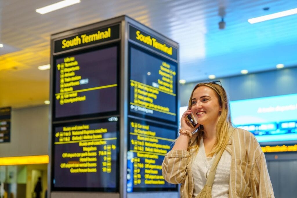 Ryanair change flight date: a traveller on the phone in an airport terminal, departure screens behind her