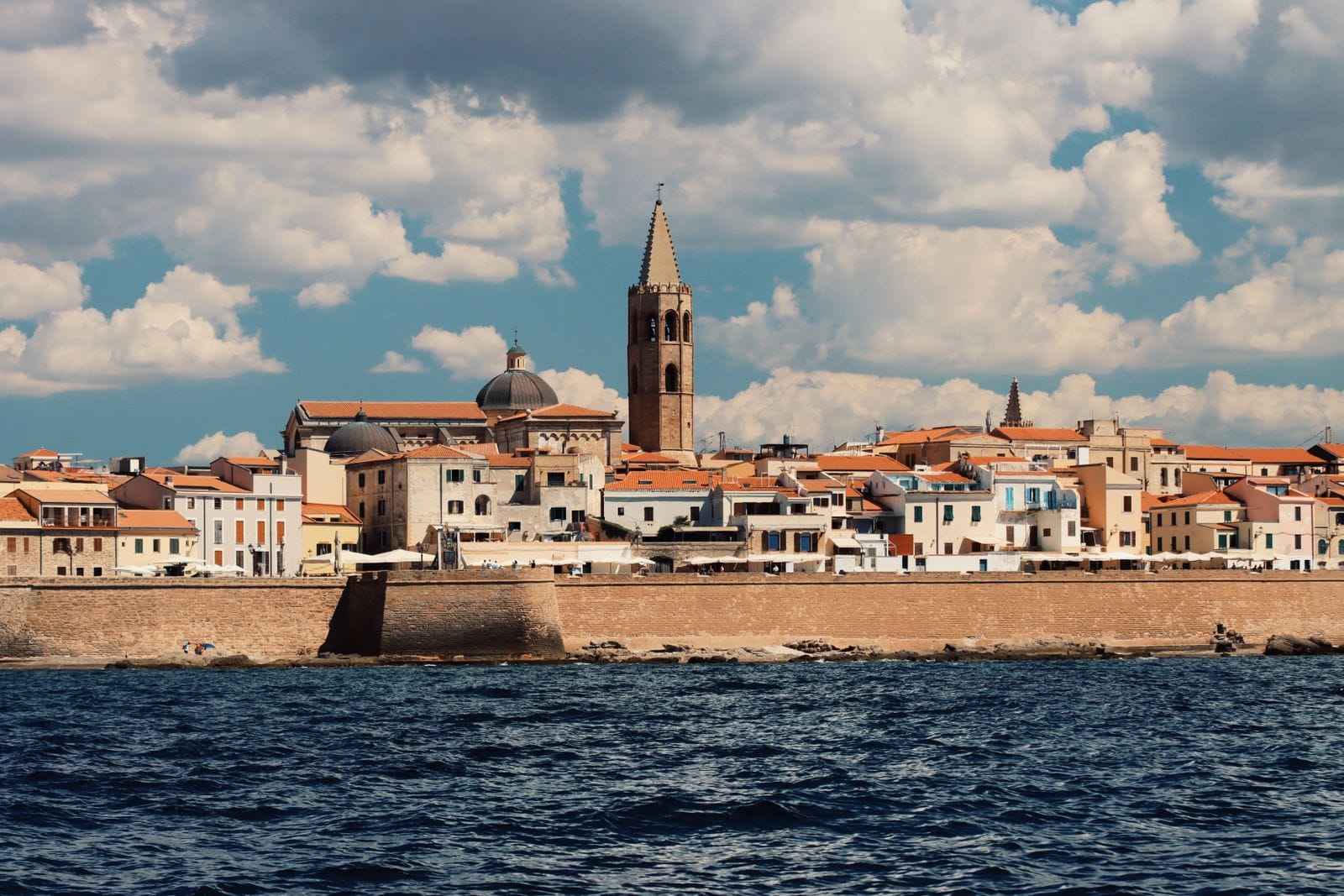 Alghero seen from the water, with the honey-coloured sea walls below the old town, terracotta roofs and the cathedral bell tower