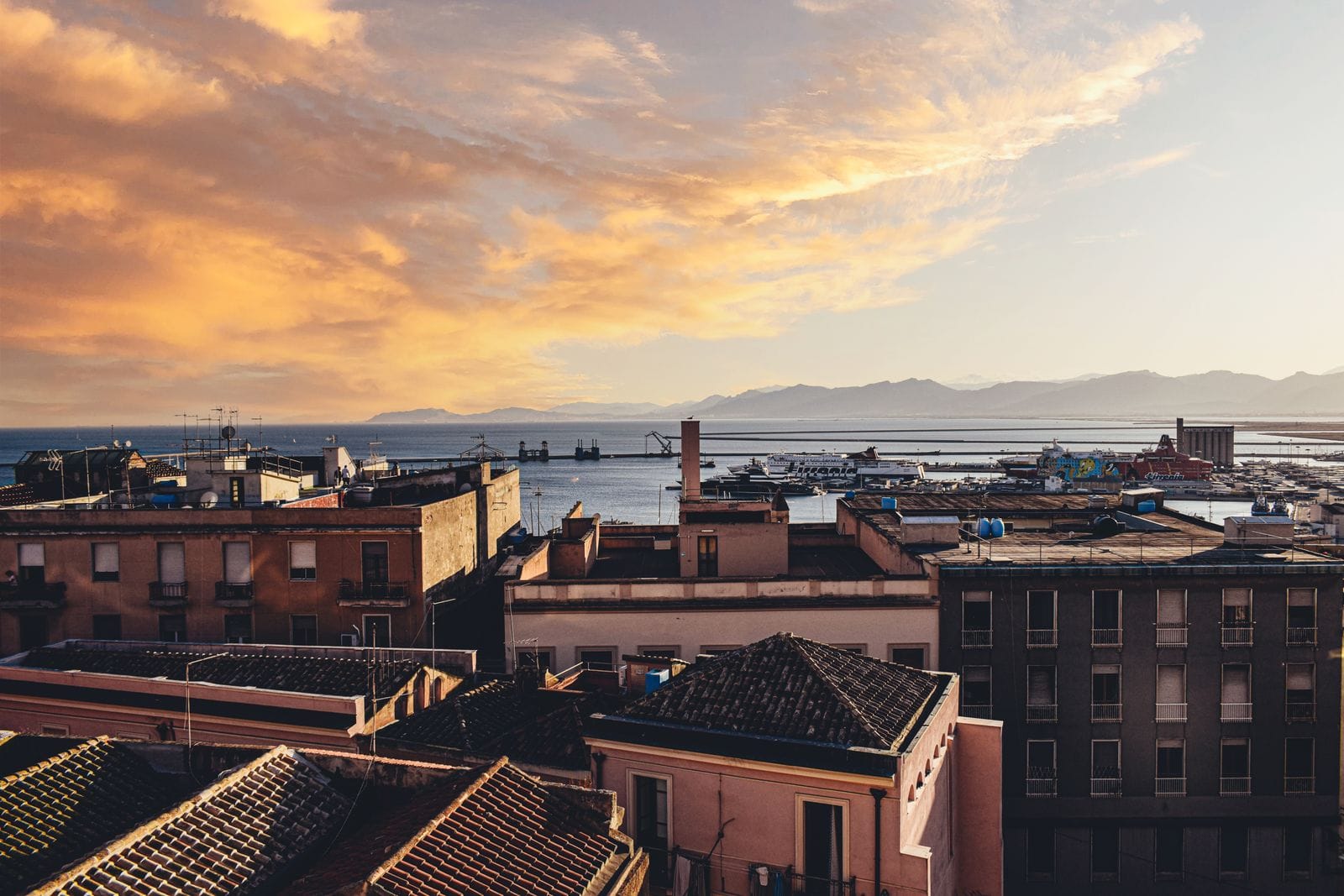 Cagliari rooftops at sunset looking down over the working port, with ferries at the quay and mountains across the gulf