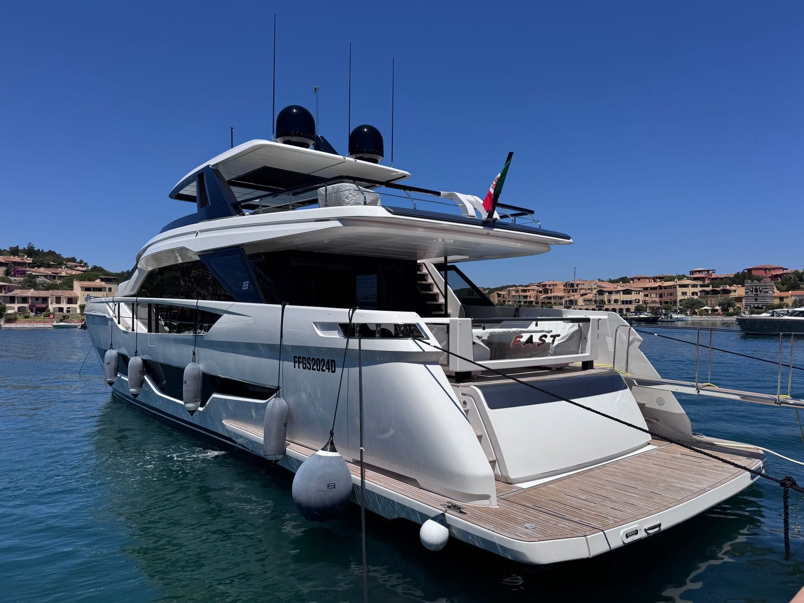 A large white motor yacht moored stern-on in a Costa Smeralda marina, with terracotta-roofed village buildings on the hillside behind