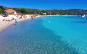 Is Sardinia expensive: bathers in clear turquoise shallows off a white sand beach in Sardinia, with low green scrub headland behind