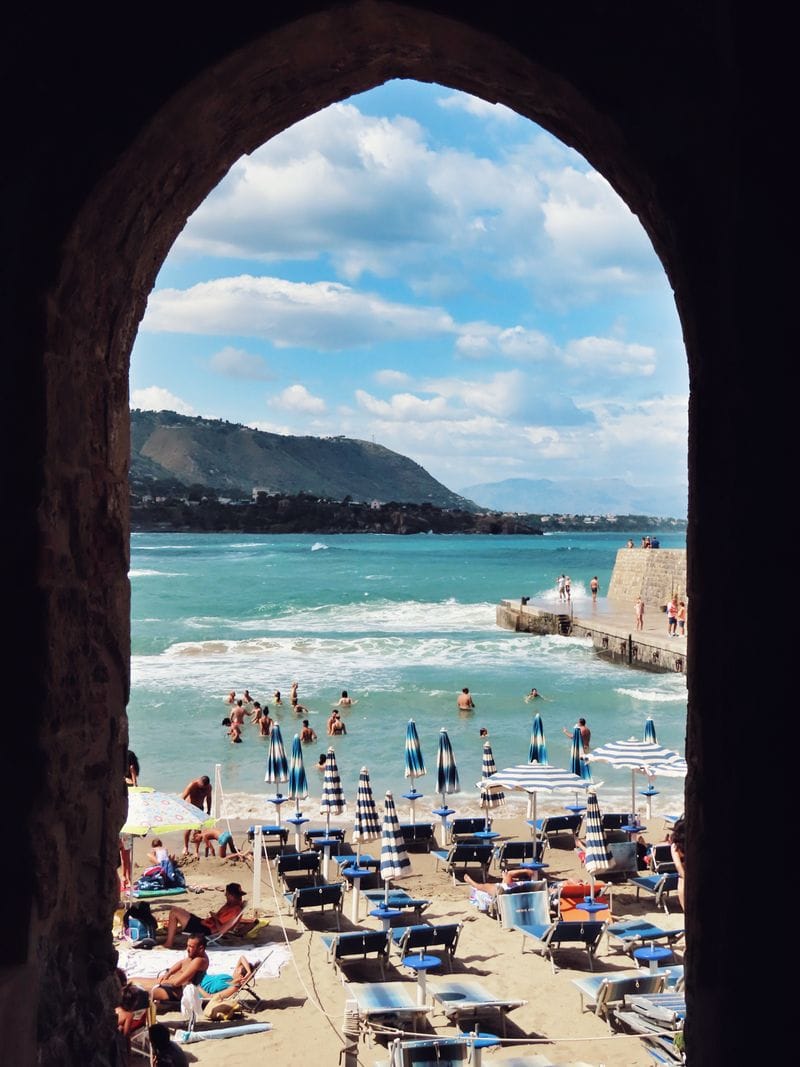 Beach umbrellas and swimmers seen through a stone archway on a Sicilian beach