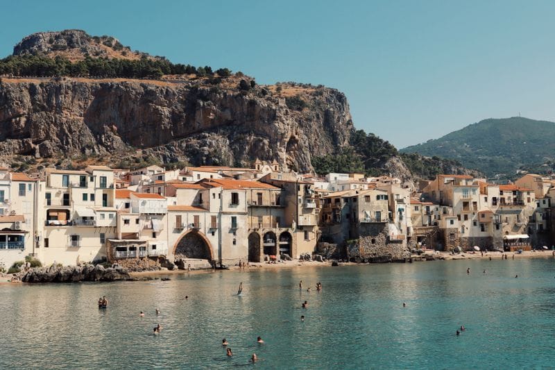 Swimmers in the clear turquoise water off Cefal&ugrave;'s beach, cliff rising behind the old town