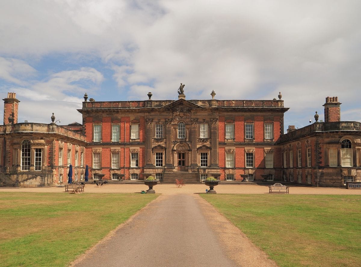 A red-brick baroque English country house with formal lawns and a gravel drive