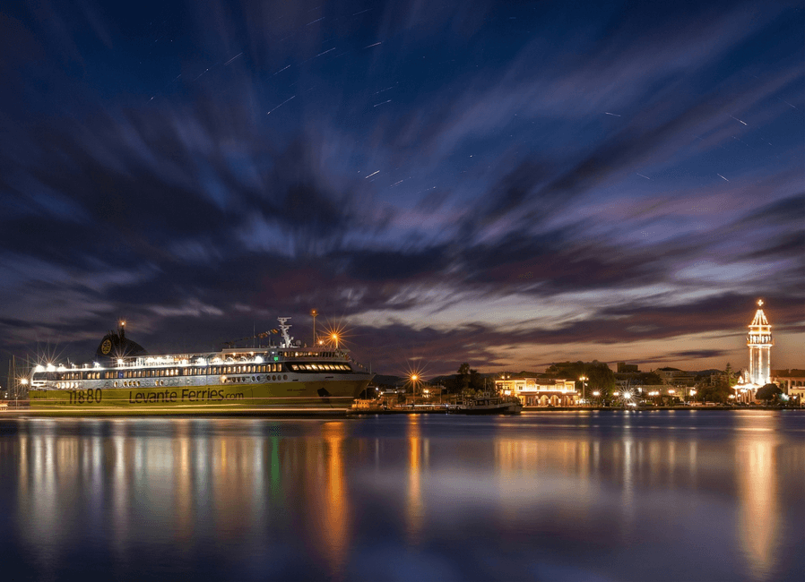 A ferry moored at Zakynthos port at night with a lighthouse and sky reflected in calm water