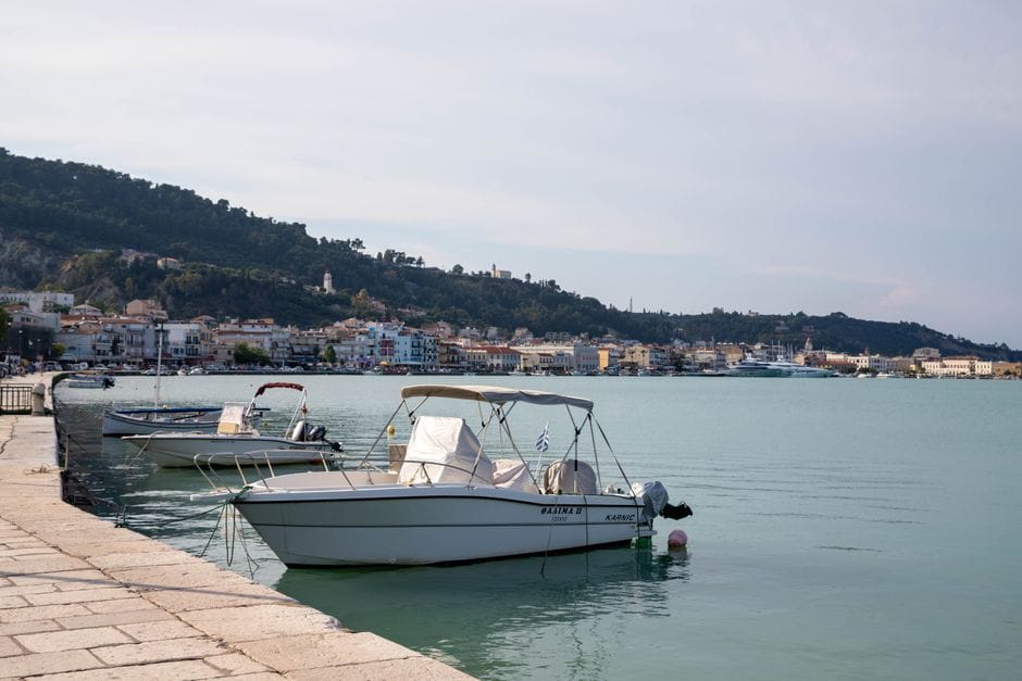 Small boats moored in Zakynthos Town harbour with the coastline and hills behind under a clear summer sky