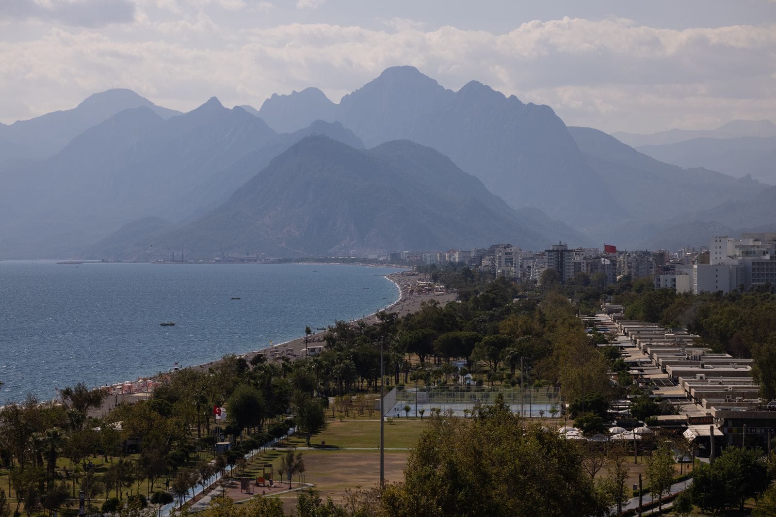 Antalya's palm-lined seafront and pebble beach curving beneath the Taurus Mountains, high-rise hotels along the shore