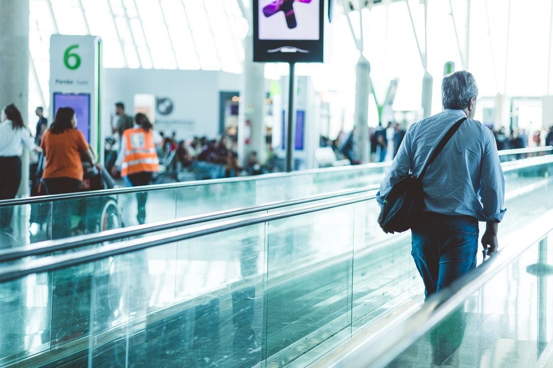 A passenger with a shoulder bag walks along a moving walkway in a bright airport terminal