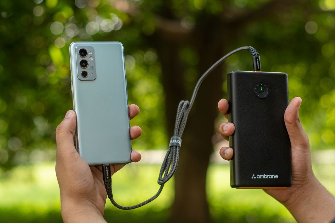 Two hands holding a smartphone connected by a braided cable to a black power bank, with trees behind