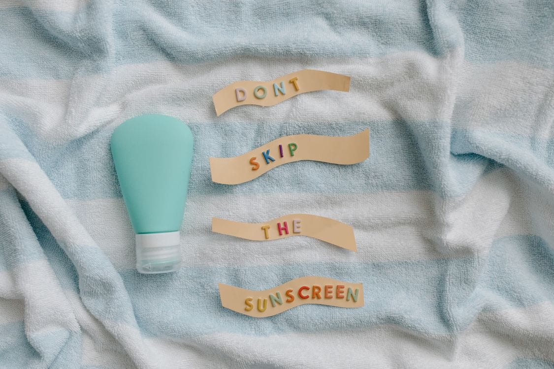 A teal travel squeeze bottle on a striped beach towel beside paper strips spelling out a sun cream reminder