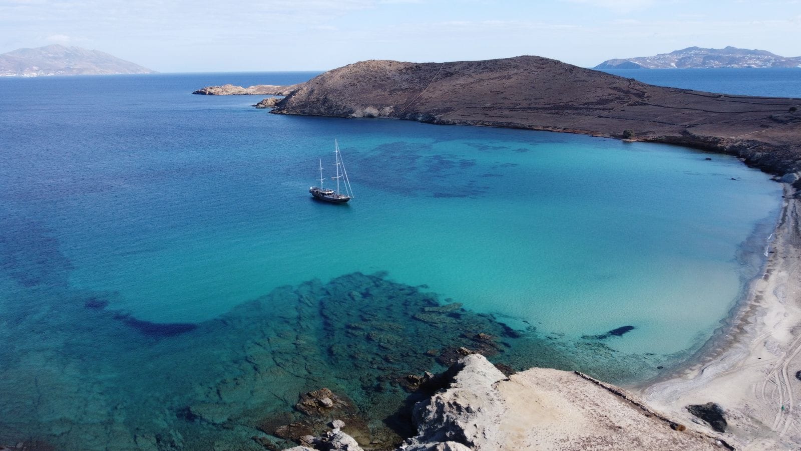 Aerial view of a quiet turquoise bay near Mykonos, Greece, with a single sailboat anchored offshore