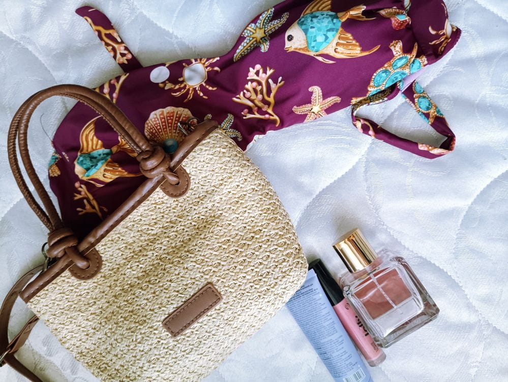 A straw beach bag, patterned swimsuit, perfume bottle and lip gloss laid out on a white bedspread