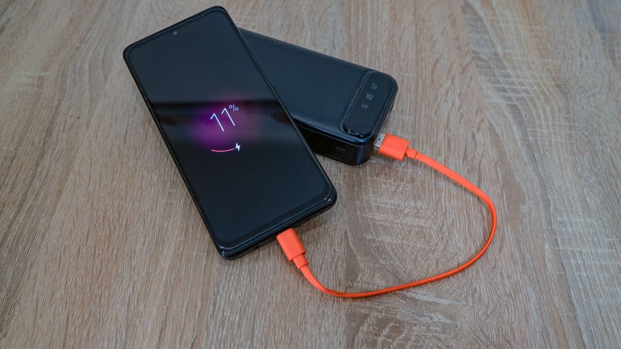 A smartphone charging from a black power bank through an orange cable on a wooden table