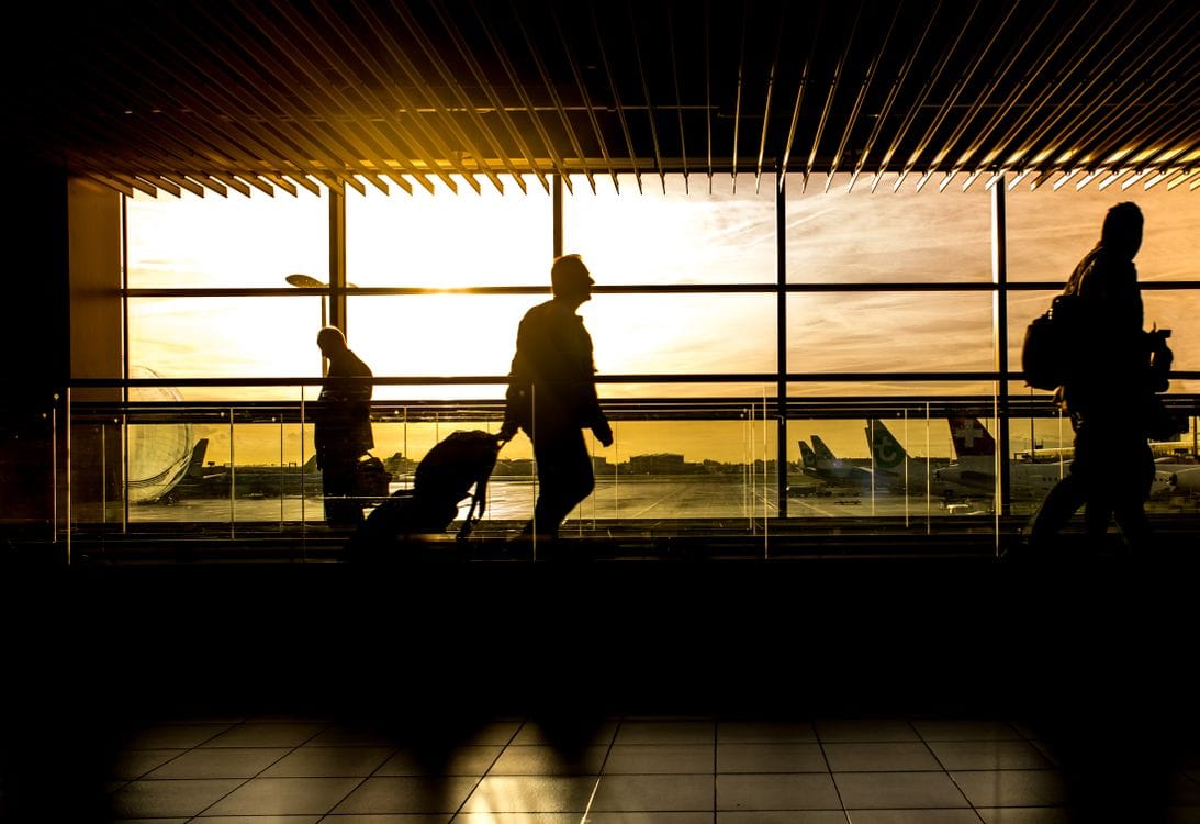 Silhouettes of travellers pulling luggage past a terminal window at sunset, with parked aircraft outside