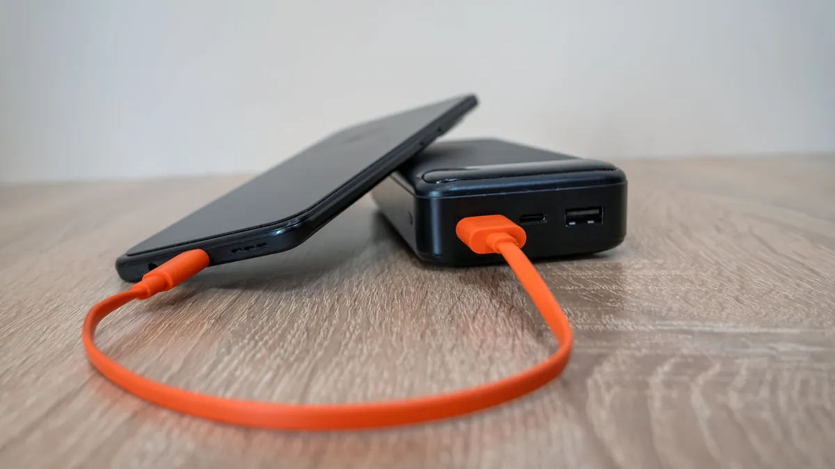 A smartphone charging from a black power bank through an orange cable on a wooden table