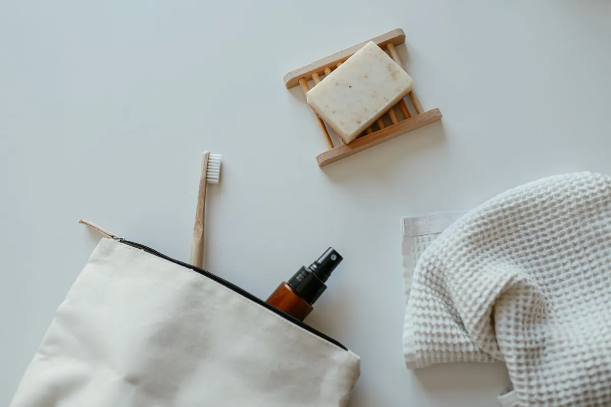 A canvas wash bag holding a spray bottle, bamboo toothbrush and bar of soap, next to a folded towel