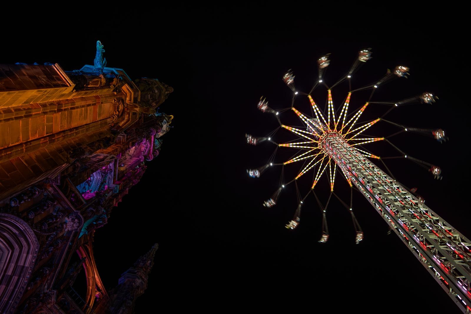 The Starflyer swing ride lit up at night beside a floodlit Gothic monument in Edinburgh