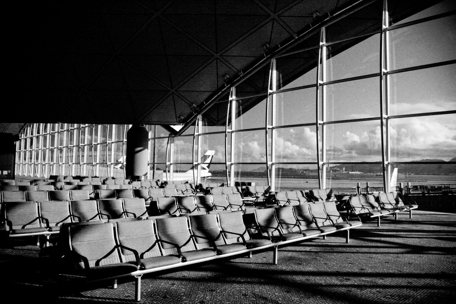 Rows of empty seats in a departure lounge in black and white, with runway views through tall windows