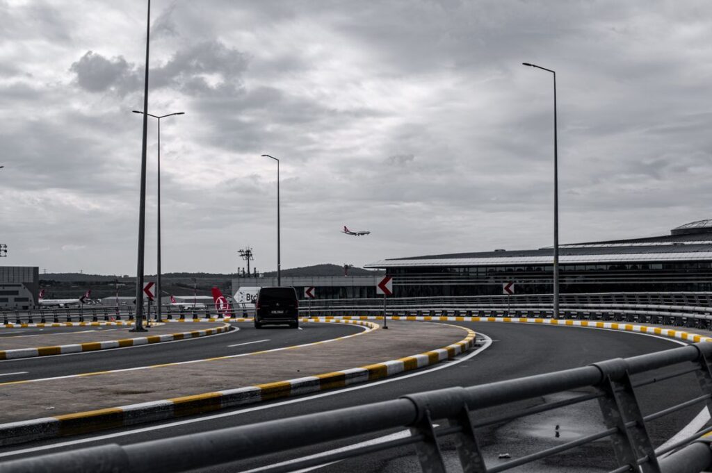 A dark people carrier on a curving airport access road under grey cloud, the approach to a terminal like the Luton airport drop off zone
