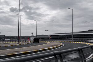 A dark people carrier on a curving airport access road under grey cloud, the approach to a terminal like the Luton airport drop off zone