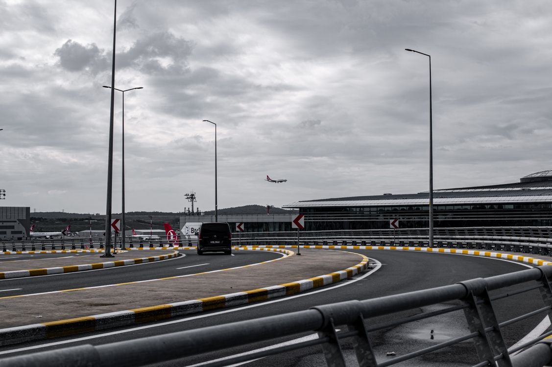 A dark people carrier on a curving airport access road under grey cloud, the approach to a terminal like the Luton airport drop off zone