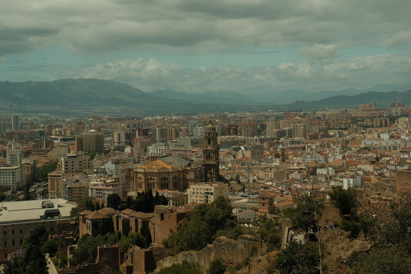 Hillside fortress ruins among trees above Malaga, with the city and mountains stretching into the distance