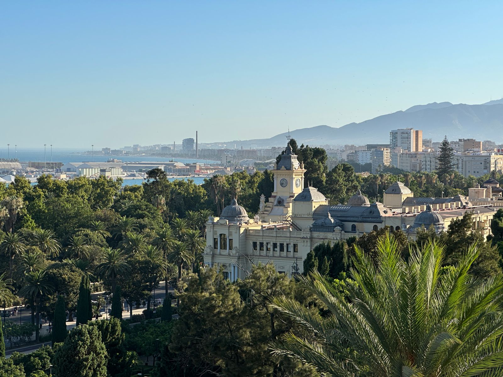 A clock-towered civic building behind palm trees and park gardens in Malaga, with the sea and mountains beyond