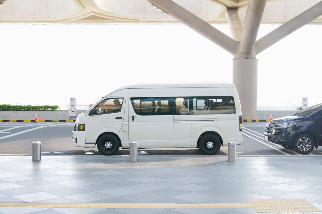 A white minivan stopped at an airport terminal drop-off zone, where Manchester Airport Terminal 2 charges by the minute