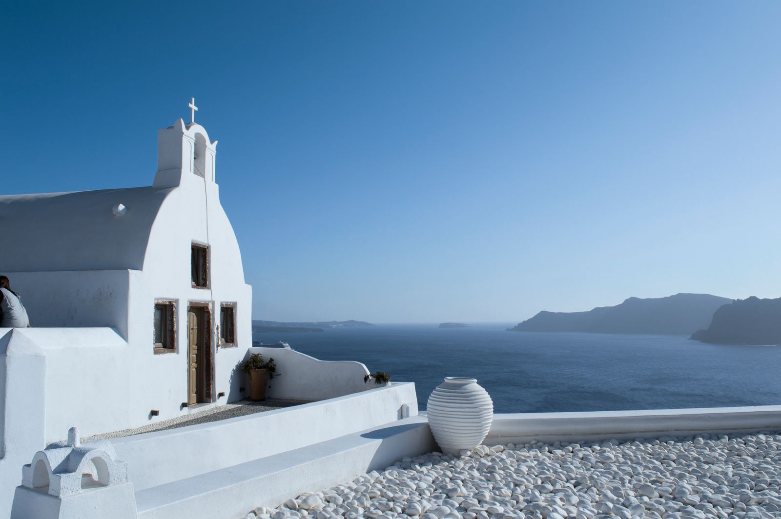 A whitewashed chapel and a white stone jar on a pebble terrace above the Aegean in Santorini